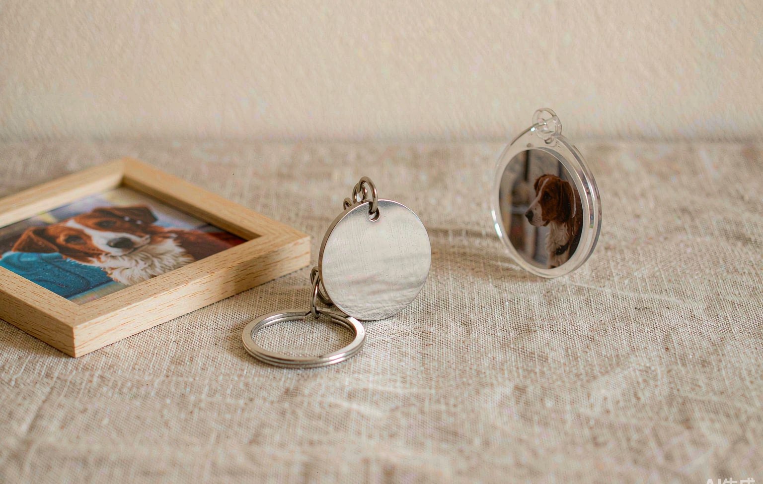 A small engraved metal pet ID tag, a photo keychain and a framed pet photo resting on a linen surface in soft daylight