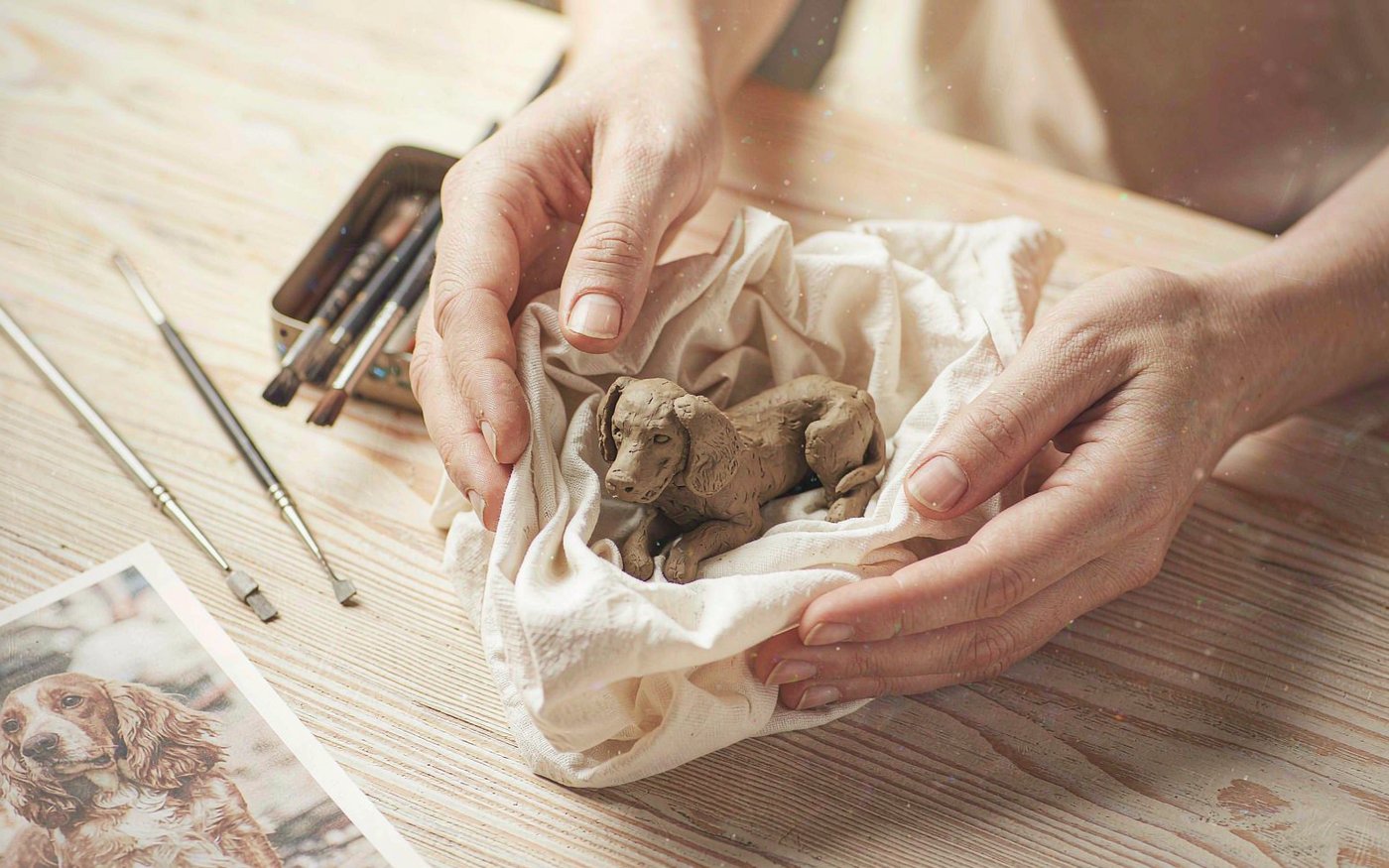 Hands wrapping a hand-sculpted clay pet figurine in tissue paper at a workshop bench beside sculpting tools and a reference photo