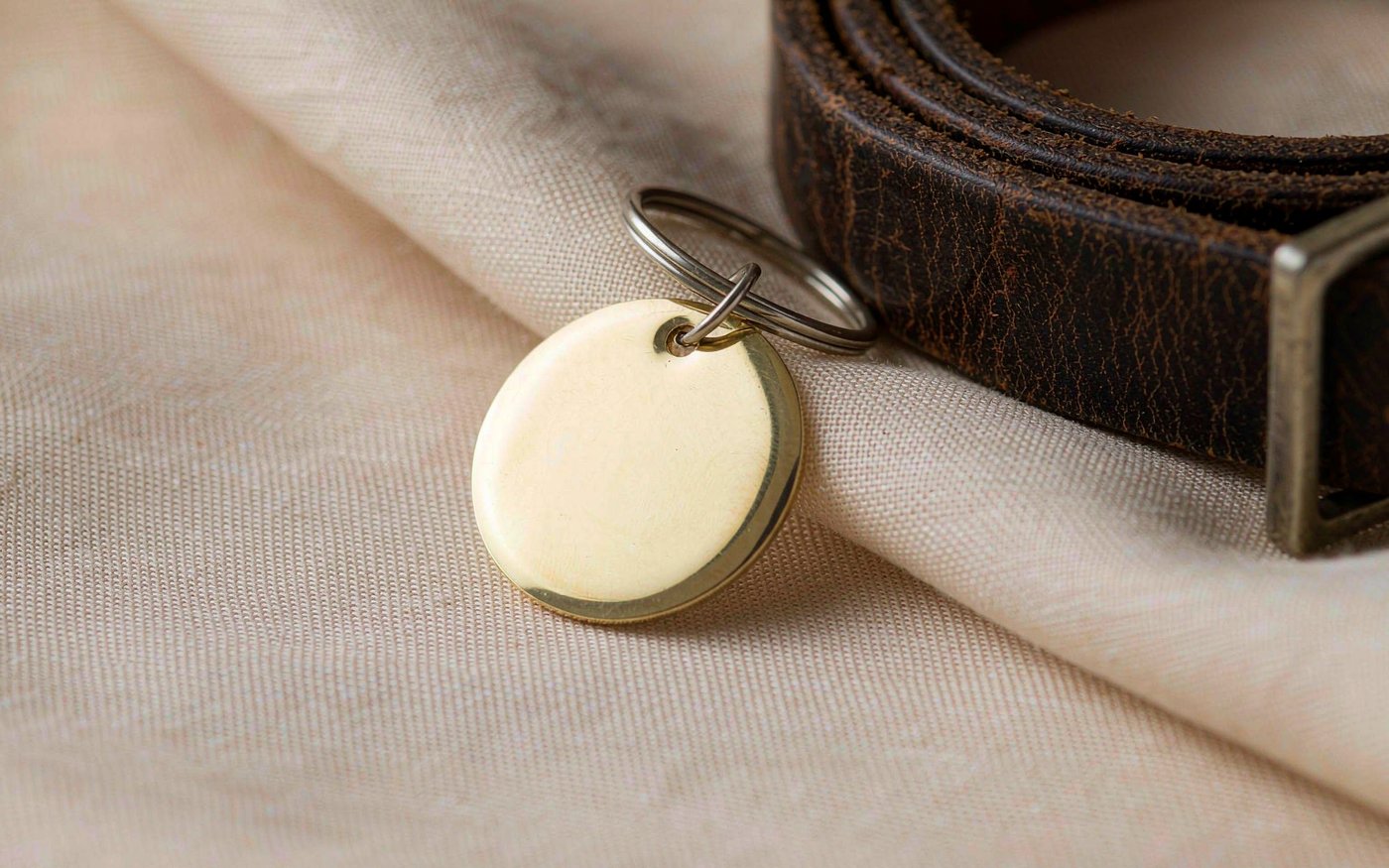 A small blank polished brass pet tag on a steel keyring resting beside a weathered leather dog collar on cream linen