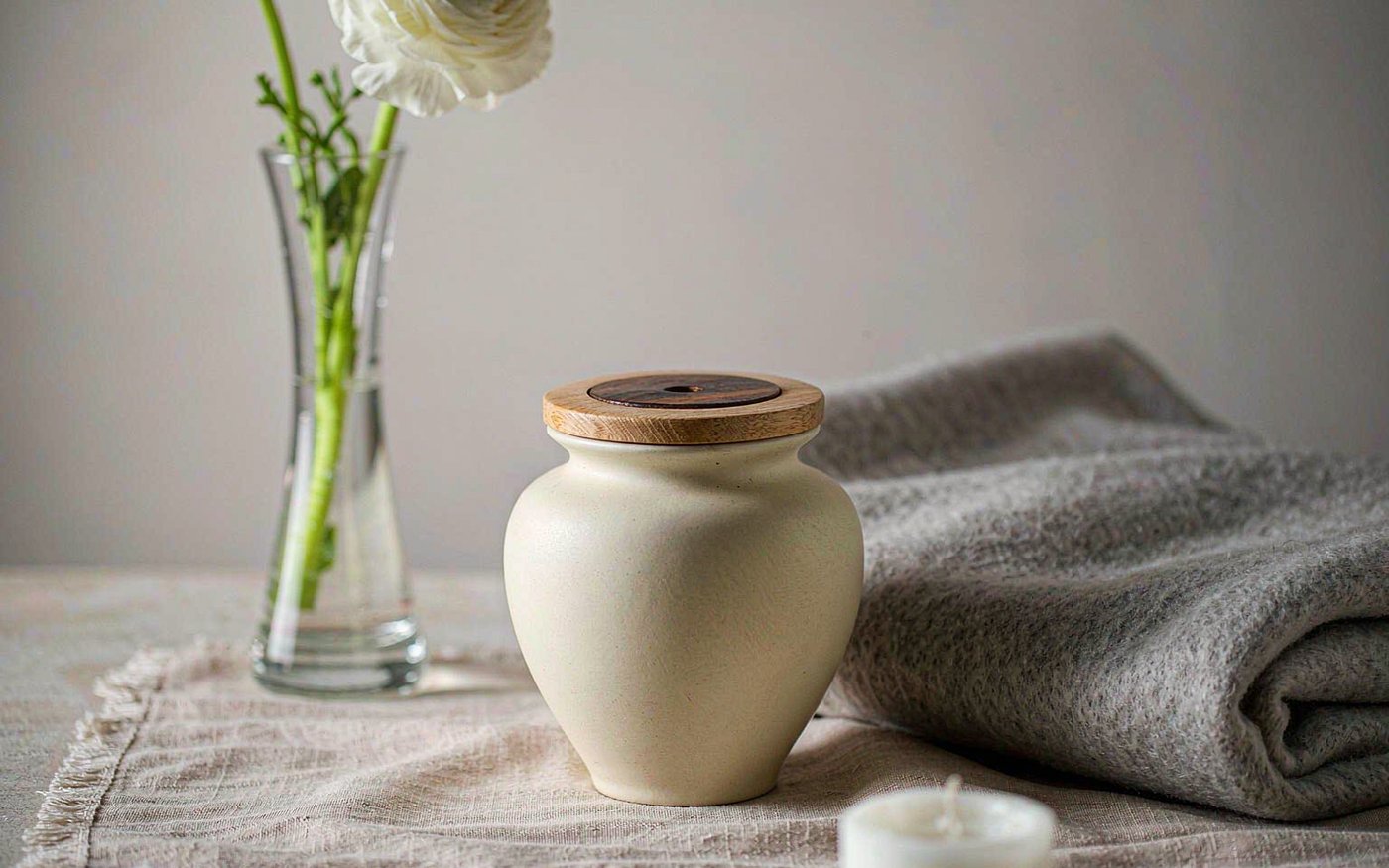 A small cream ceramic memorial urn with a wooden lid beside a white flower in a glass vase, a folded grey blanket and an unlit candle on a linen runner