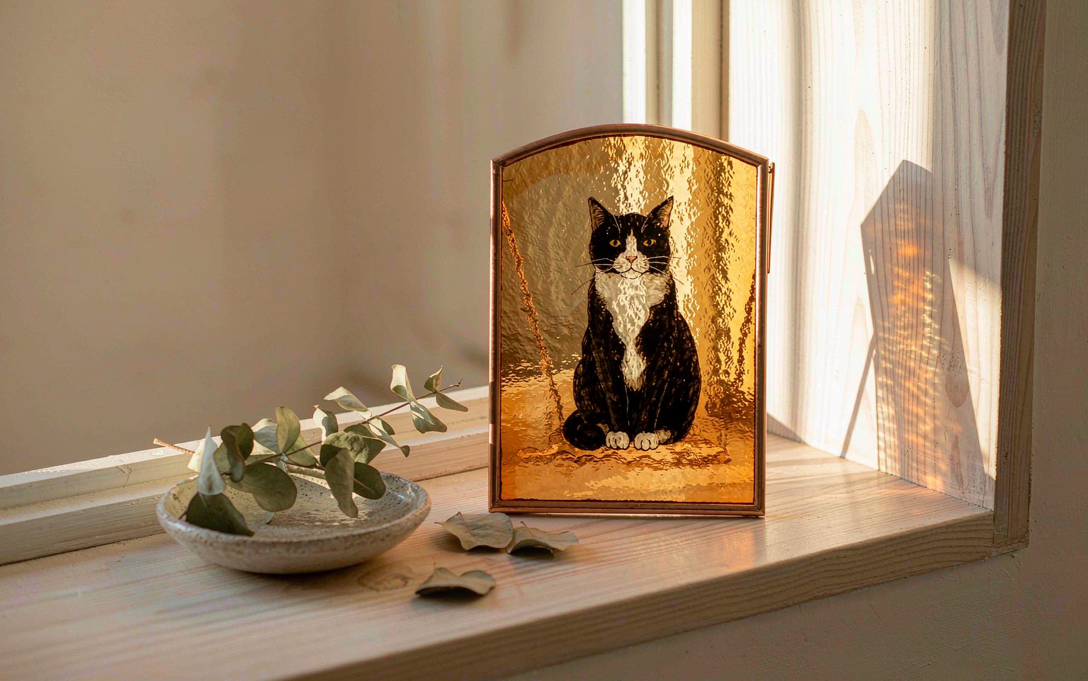 A hand-painted glass cat portrait in a copper frame on a sunlit windowsill — a quiet pet loss gift for a cat lover