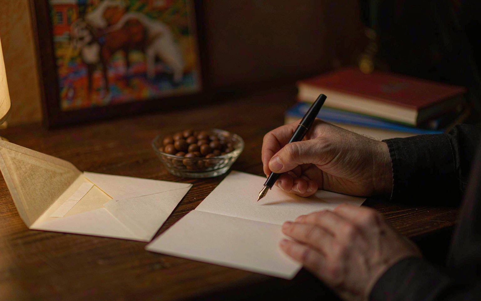 A person's hands writing a message in a sympathy card at a wooden desk with an open envelope and a framed pet photograph nearby